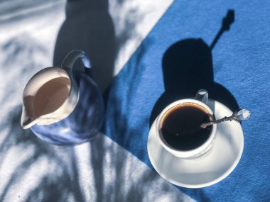 Beyaz bardakta kahve, bir kaşık ve küçük bir sürahi süt, mavi ve beyaz arka planda, kuru yabani çimen gölgeleri.