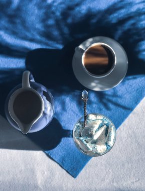 Beyaz bardakta kahve, bardakta dondurma mavi ve beyaz arka planda küçük bir sürahi süt ve kuru yabani çimen gölgeleri.