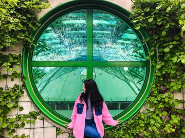 Varşova Üniversitesi Kütüphanesi 'nin çatısındaki pembe ceketli genç bayan yeşil sarmaşık çalılıklarının kenarındaki yeşil yuvarlak pencerenin yanında.