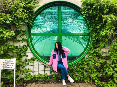 Varşova Üniversitesi Kütüphanesi 'nin çatısındaki pembe ceketli genç bayan yeşil sarmaşık çalılıklarının kenarındaki yeşil yuvarlak pencerenin yanında.