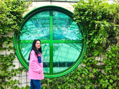 Varşova Üniversitesi Kütüphanesi 'nin çatısındaki pembe ceketli genç bayan yeşil sarmaşık çalılıklarının kenarındaki yeşil yuvarlak pencerenin yanında.