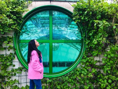 Varşova Üniversitesi Kütüphanesi 'nin çatısındaki pembe ceketli genç bayan yeşil sarmaşık çalılıklarının kenarındaki yeşil yuvarlak pencerenin yanında.