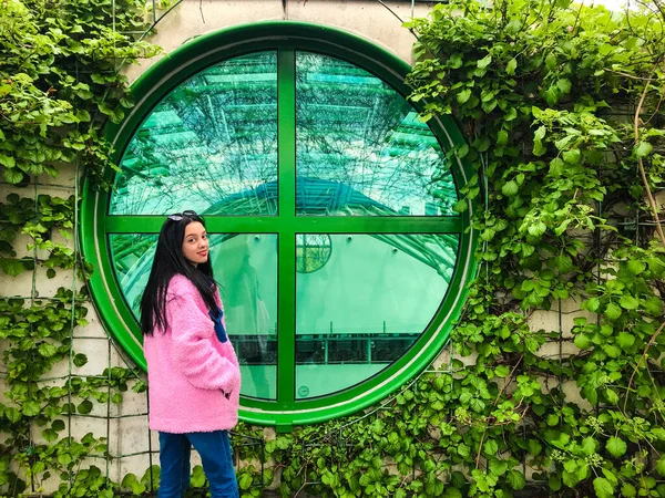 Varşova Üniversitesi Kütüphanesi 'nin çatısındaki pembe ceketli genç bayan yeşil sarmaşık çalılıklarının kenarındaki yeşil yuvarlak pencerenin yanında.
