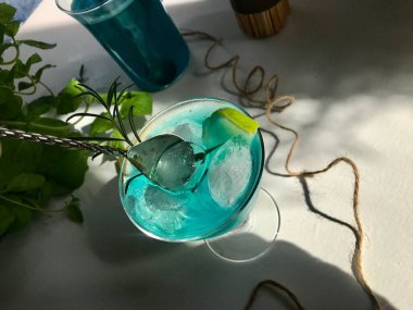 Mavi buzlu bir bardakta mavi bir içecek. Mavi arka planda beyaz masada buz küpleri var. Ayrıca açık pencereden doğal güneş ışığı alan nane yaprağı, biberiye, ip ve kaşık kullanılmış.