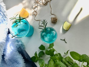 Mavi buzlu bir bardakta mavi bir içecek. Mavi arka planda beyaz masada buz küpleri var. Ayrıca açık pencereden doğal güneş ışığı alan nane yaprağı, biberiye, ip ve kaşık kullanılmış.