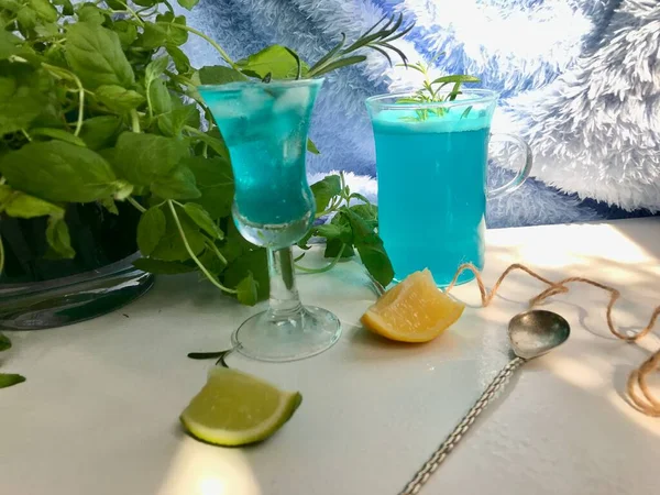 青い背景の白い机の上に氷のキューブとガラスの中の青い冷たい飲み物 使用ミントの葉とローズマリー ロープ オープンウィンドウからの自然な日光とスプーン — ストック写真