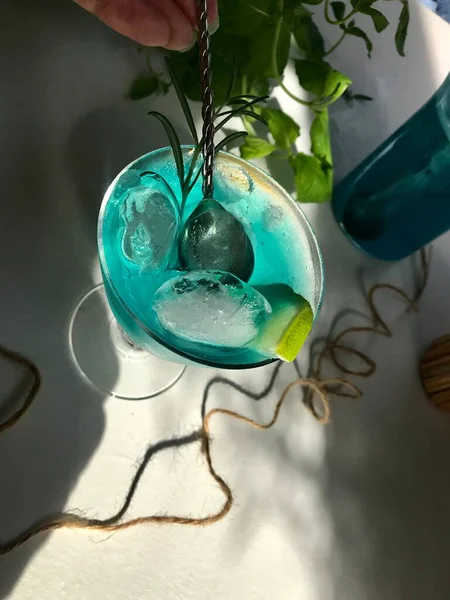 青い背景の白い机の上に氷のキューブとガラスの中の青い冷たい飲み物 使用ミントの葉とローズマリー ロープ オープンウィンドウからの自然な日光とスプーン — ストック写真