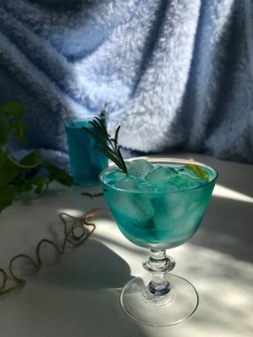 Mavi buzlu bir bardakta mavi bir içecek. Mavi arka planda beyaz masada buz küpleri var. Ayrıca açık pencereden doğal güneş ışığı alan nane yaprağı, biberiye, ip ve kaşık kullanılmış.