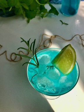 Mavi buzlu bir bardakta mavi bir içecek. Mavi arka planda beyaz masada buz küpleri var. Ayrıca açık pencereden doğal güneş ışığı alan nane yaprağı, biberiye, ip ve kaşık kullanılmış.