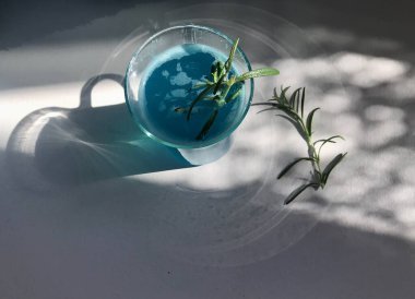 Mavi buzlu bir bardakta mavi bir içecek. Mavi arka planda beyaz masada buz küpleri var. Ayrıca açık pencereden doğal güneş ışığı alan nane yaprağı, biberiye, ip ve kaşık kullanılmış.