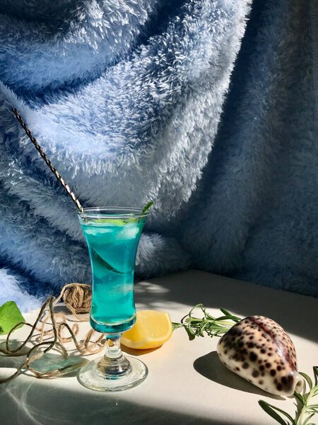 Голубой холодный напиток в стакане с кубиками льда на белом столе с голубым фоном. Также используются мятные листья и розмарин, веревка, ложка с натуальным солнечным светом из открытого окна