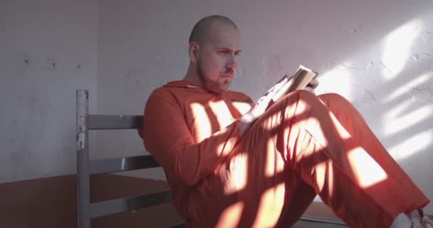 Hapishane hücresinde kitap okuyan turuncu tulumlu bir mahkum yatakta oturuyor.. — Stok video