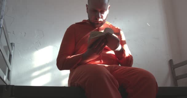 Узник в оранжевом комбинезоне читает книгу в тюремной камере, сидящей на кровати. — стоковое видео