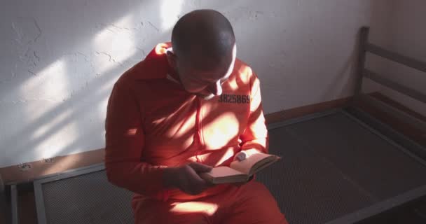刑務所で読書する オレンジ色の制服の囚人が 刑務所の刑務所で本を読んでる 囚人は祈りの本を読み バーから窓を眺める 刑務所における宗教 — ストック動画