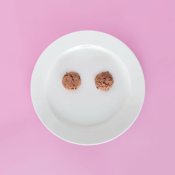 Happy face emoji made of wholemeal delicious cookies in white plate on isolated pastel pink background. Top view. Aesthetic abstract concept of weight loss dieting or healyhy wholegrain food. Health benefits of eating whole grains. Minimal flat lay.