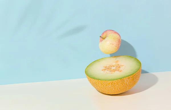 Half of juicy fresh melon, apple and shadow of tropical palm on isolated pastel beige and blue background. Minimal summer fruit scene. Raw food concept. Abstract creative idea of vacation on the see.