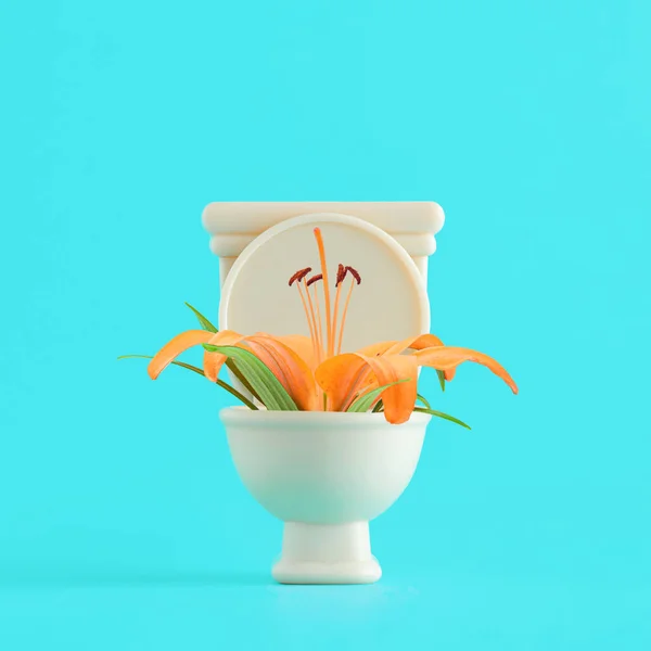 Orange lily flower with green leaves in beige toilet on isolated vibrant, turquoise-blue background. Minimal spring or summer floral, optimistic, hopefulness concept. The idea of the bright side of life, cleanliness or good digestion.