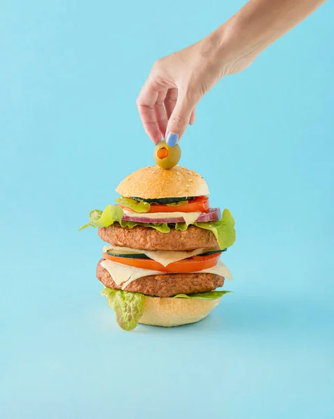 A woman\'s hand puts an olive on bun with pea protein veggie burger, lettuce, tomato, cheese, onion and cucumber. Isolated pastel blue background. Concept of healthy or unhealthy diet or food overdose.