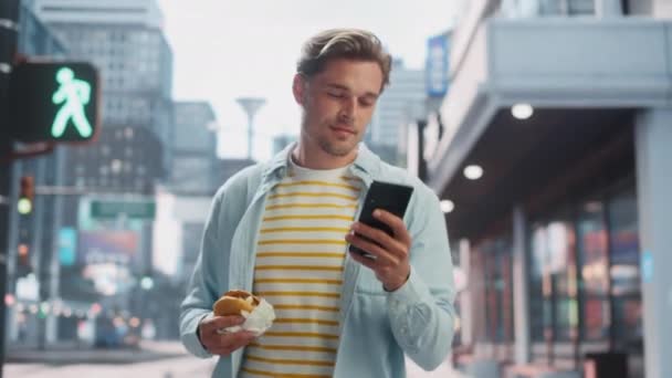 Hombre guapo comiendo hamburguesa y el uso de Smartphone en la ciudad — Vídeos de Stock