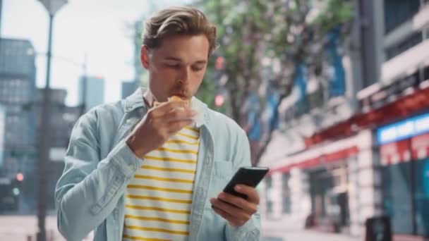 Hombre guapo comiendo hamburguesa y el uso de Smartphone en la ciudad — Vídeos de Stock