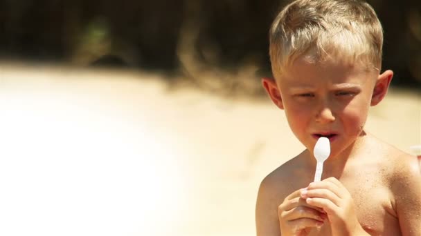 小さな男の子は、日当たりの良いビーチでヨーグルトを食べる — ストック動画