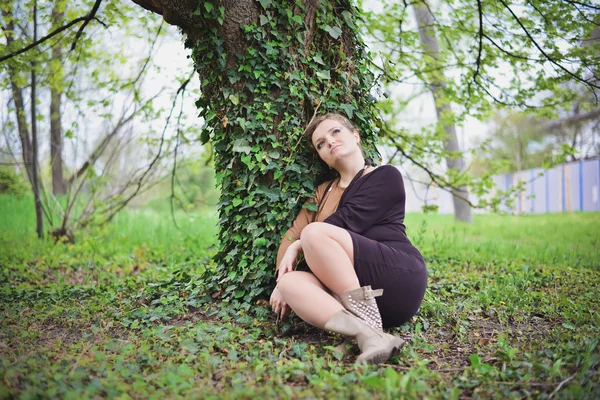 Girl in black and brown dress sits near a tree — Stock Photo, Image
