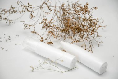 Üzerinde yazı olmayan kozmetik, hijyen veya tıbbi ürünler için iki beyaz şişe veya tüp, kurumuş çiçekli hafif bir arkaplan üzerine yazı.