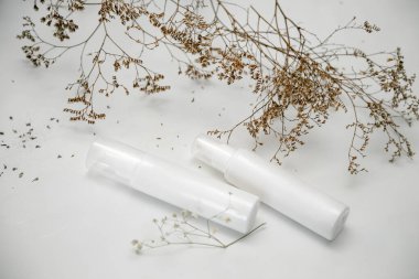 Üzerinde yazı olmayan kozmetik, hijyen veya tıbbi ürünler için iki beyaz şişe veya tüp, kurumuş çiçekli hafif bir arkaplan üzerine yazı.