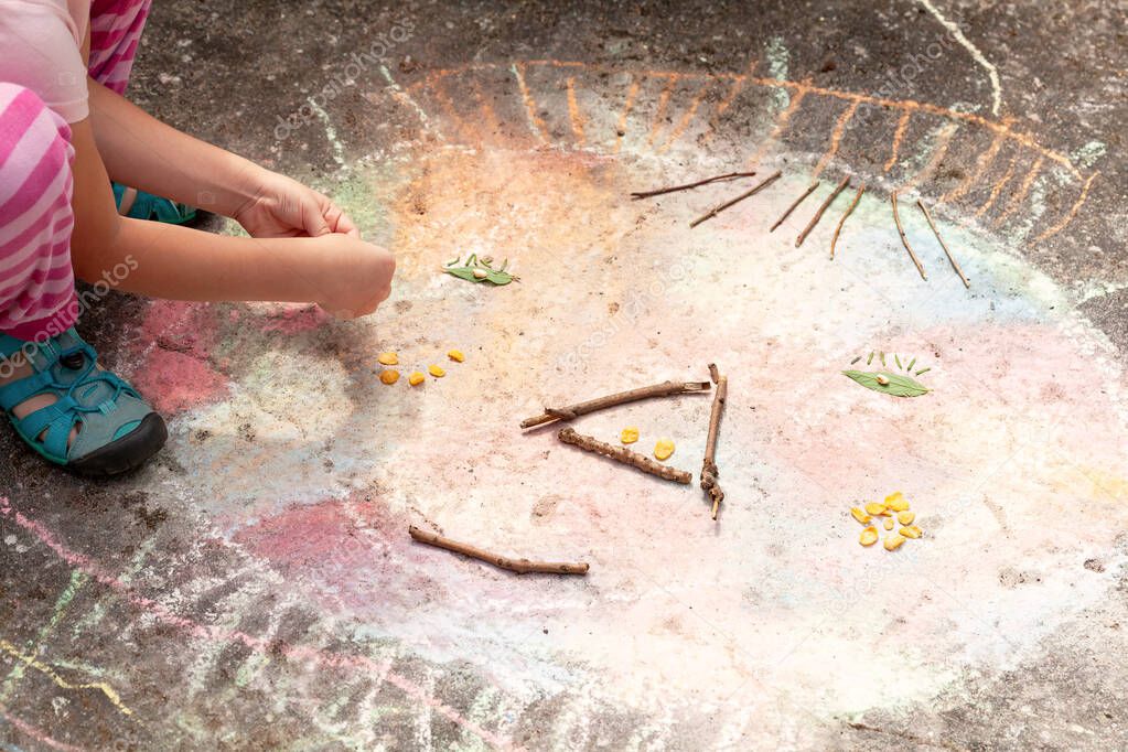 Anonymous young elementary school age child, little girl playing outside, outdoors creating a face shape from sticks, leaves and chalk. Children art therapy and sensory integration simple concept