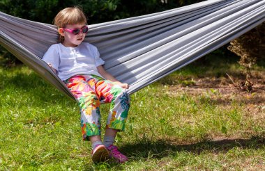 Tek bir okul çağındaki kız, güneş gözlüklü ve renkli elbiseli çocuk dışarıda, açık havada, arka bahçede bir hamakta dinleniyor. Havalı çocuk, yaz tatili boş zaman, dinlenme konsepti, fotokopi alanı, bir kişi