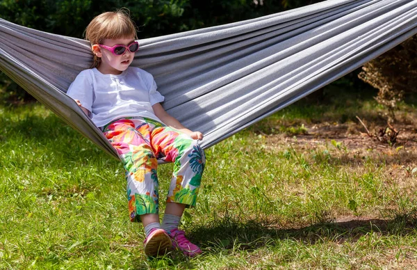 Tek bir okul çağındaki kız, güneş gözlüklü ve renkli elbiseli çocuk dışarıda, açık havada, arka bahçede bir hamakta dinleniyor. Havalı çocuk, yaz tatili boş zaman, dinlenme konsepti, fotokopi alanı, bir kişi