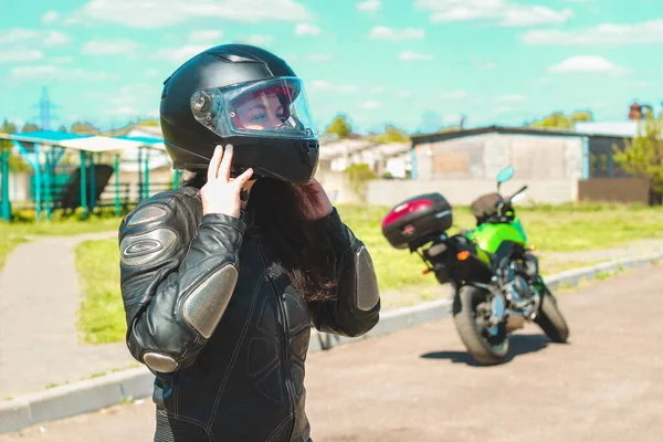 一个穿着保护摩托车皮衣的女孩在摩托车的背景下戴上头盔 — 图库照片