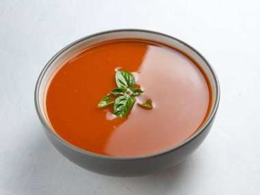 Fesleğenli domates çorbası gri bir kasede, beyaz bir surfac üzerinde.