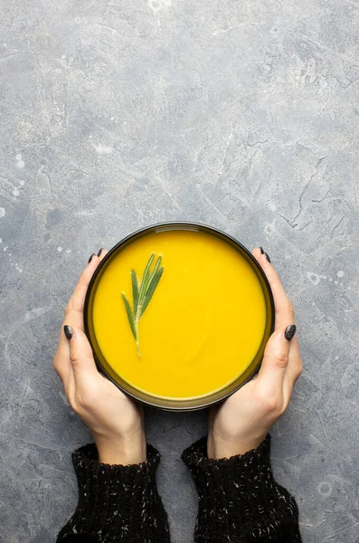 Pumpkin soup in female hands on a gray concrete background with copy space. Hands of a girl in a black sweater holding black bowl, a sprig of rosemary in the soup