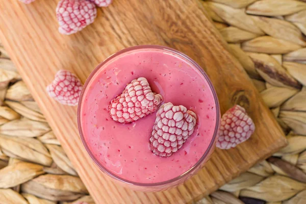 Top view of fresh raspberry smoothie in a glass on a wooden serving board. Summer refreshing berry drink with two frozen raspberries on top