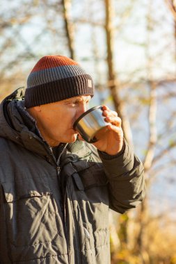 Yaşlı bir adam gölün kıyısındaki termostan sıcak çay içiyor ve arka planda renkli bir sonbahar havası var. Doğada sıcak kahve içilir. Sonbahar havası. Sonbaharda insanlar.
