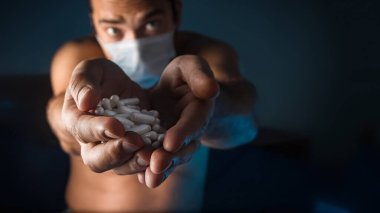 Tıbbi maskeli bronzlaşmış bir adam. Elimde beyaz haplar var. Koronavirüs salgını. Bir adamın elindeki ilaçlar..