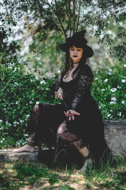 Güzel, genç, gotik, seksi ve cadı kadın. Tarzlanmış elleri, siyah tırnakları ve aksesuarları, sihirli ahşap değneği ve doğanın arasındaki ormanda cadı şapkası olan kadın.