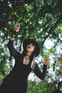 Güzel, genç, gotik, seksi ve cadı kadın. Tarzlanmış elleri, siyah tırnakları ve aksesuarları, sihirli ahşap değneği ve doğanın arasındaki ormanda cadı şapkası olan kadın.