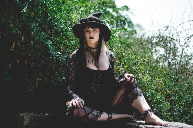 Güzel, genç, gotik, seksi ve cadı kadın. Tarzlanmış elleri, siyah tırnakları ve aksesuarları, sihirli ahşap değneği ve doğanın arasındaki ormanda cadı şapkası olan kadın.