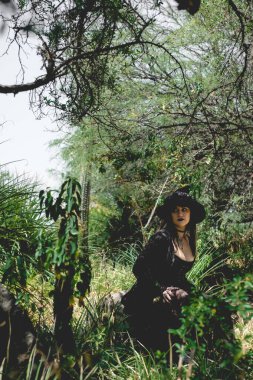 Güzel, genç, gotik, seksi ve cadı kadın. Tarzlanmış elleri, siyah tırnakları ve aksesuarları, sihirli ahşap değneği ve doğanın arasındaki ormanda cadı şapkası olan kadın.