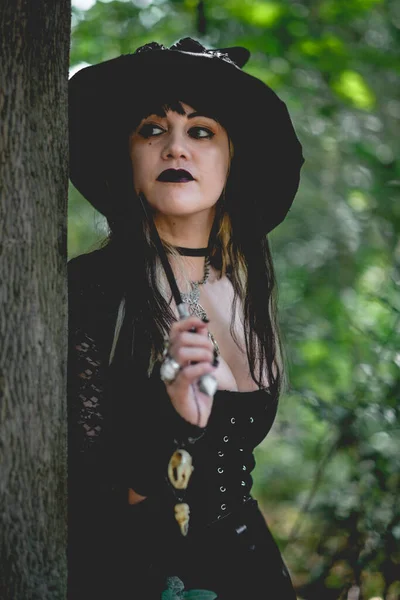 Beautiful young, gothic, sexy and witch woman from with stylized hands with black nails and accessories, magic wooden wand and witch hat in the forest between the nature