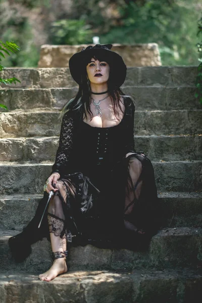 Beautiful young, gothic, sexy and witch woman from with stylized hands with black nails and accessories, magic wooden wand and witch hat in the forest in a stone stairs