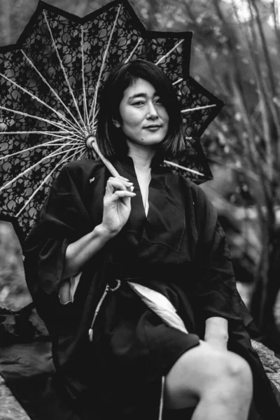 Portrait of sexy and young japanese woman with beautiful old traditional black kimono and lace sun umbrella showing legs in the autumn forest (in black and white)