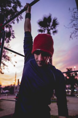 Formda ve güzel ve renkli günbatımı gökyüzü olan bir sokak çalışma parkında spor yapan, yün şapkalı, spor yapan yakışıklı, atletik Latin bir adam.