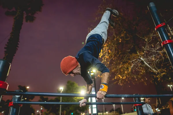 健康的年轻拉丁男子 头戴羊毛帽 在一个阳光灿烂 色彩斑斓的街头健身公园里做健美操 在高杠上倒立 — 图库照片