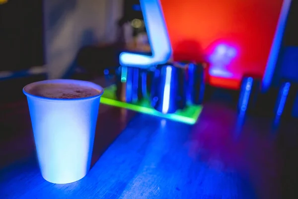 White paper cup with coffee, vintage coffee machine and coffee barista tools and jugs over a neon green tray in a coffee shop interior in the night with dark blue lights