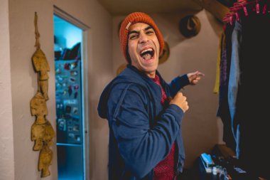 Gülümseyen ve gülen kapüşonlu ve örgü ören turuncu yün şapkalı yakışıklı Latin adam evde duruyor.