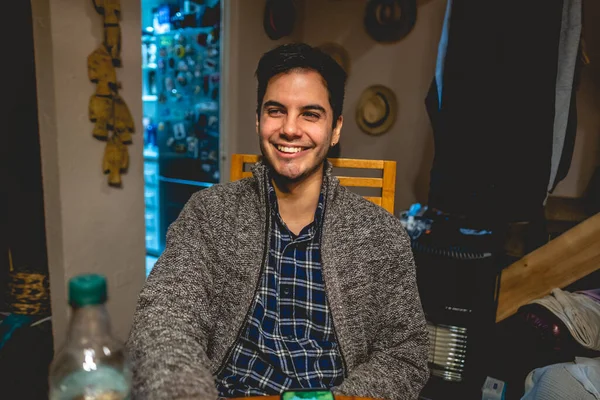Smiling and laughing young handsome latin man with beard, shirt and sweater seated in the table in home