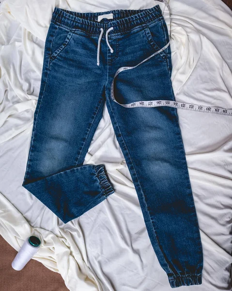 Fashion blue jeans over wrinkled white sheet with vinyl measure tape and thread cones